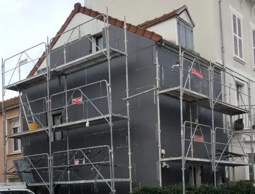 Une maison de deux étages avec un toit en pente est recouverte d'un échafaudage pour la construction ou la rénovation. Le bâtiment, qui fait partie d'un chantier d'isolation thermique (ITE), est recouvert de panneaux isolants.