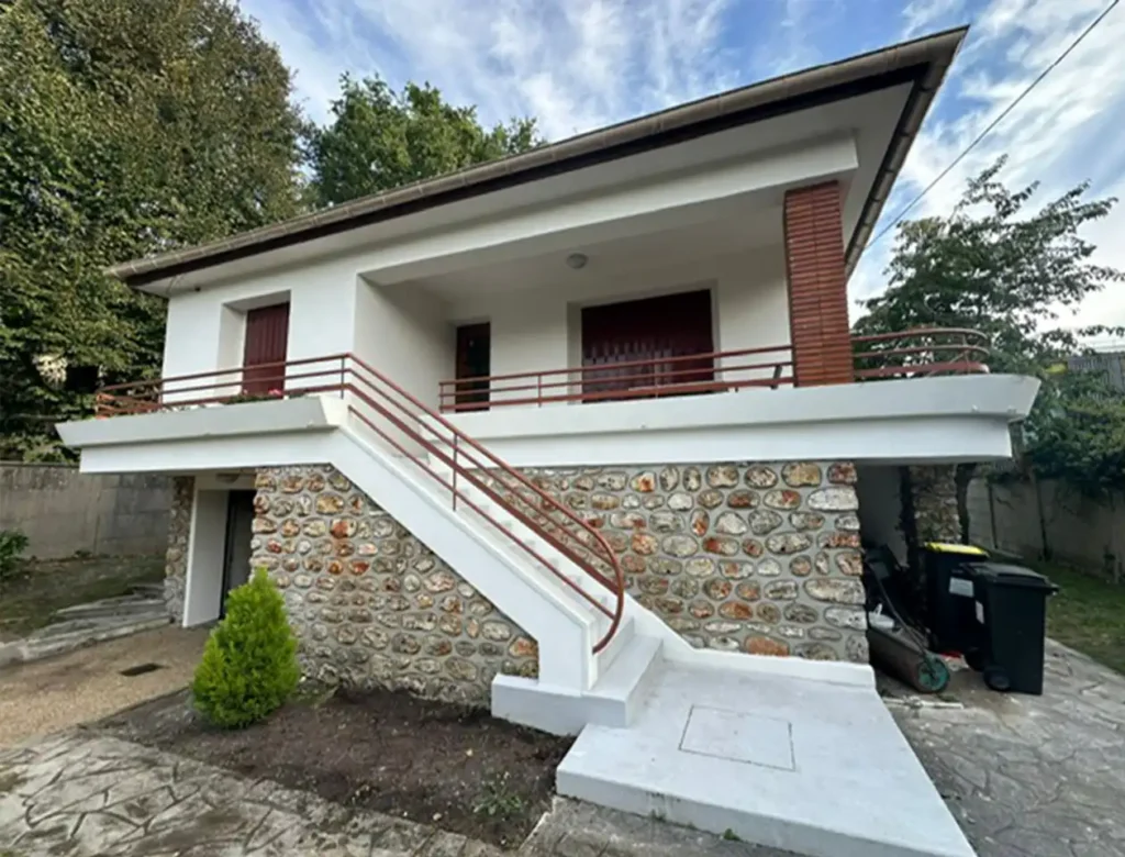 Une maison blanche avec une façade inférieure en pierre, des grilles rouges et un escalier menant à un porche couvert. Des arbres et de la verdure entourent la maison, où un chantier de rénovation ITE est en cours. Une poubelle est visible sur le côté droit sous un ciel partiellement nuageux.