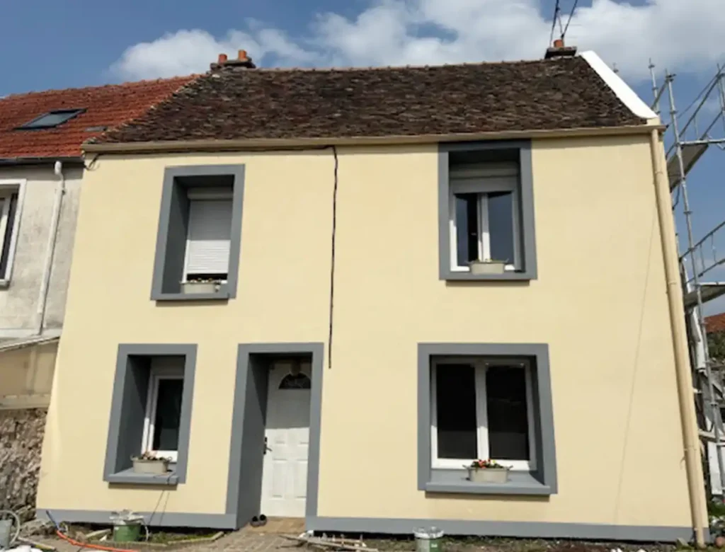 Une petite maison beige de deux &eacute;tages avec un toit en tuiles brunes, des cadres de fen&ecirc;tres gris et une porte blanche. Malgr&eacute; la fin de chantier d'un ravalement, une fissure visible court encore au centre de la fa&ccedil;ade. Quatre fen&ecirc;tres sont orn&eacute;es de charmantes jardini&egrave;res.