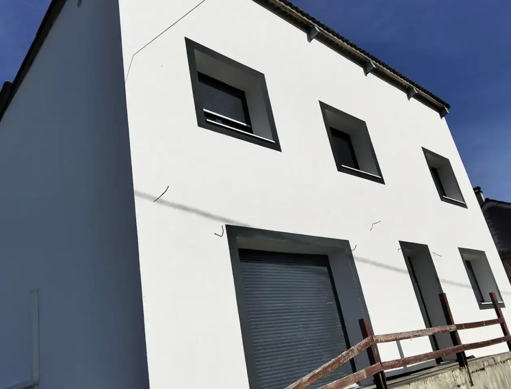 Bâtiment moderne blanc avec trois fenêtres rectangulaires et un grand volet roulant gris au rez-de-chaussée. Une clôture en bois longe la façade, mettant en valeur le ravalement de façade frais sous un ciel bleu clair.