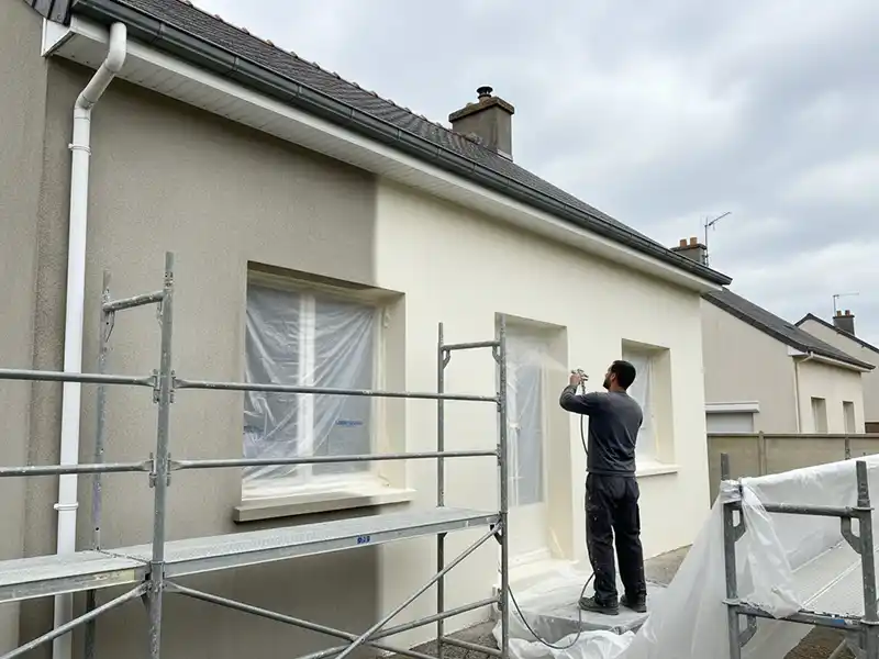 Peinture de façade - Isolex Une personne utilise un pistolet à peinture pour peindre une façade, c'est-à-dire peindre le mur extérieur d'une maison en blanc. Les fenêtres sont recouvertes de feuilles de plastique pour les protéger et un échafaudage est installé à proximité par une journée nuageuse.