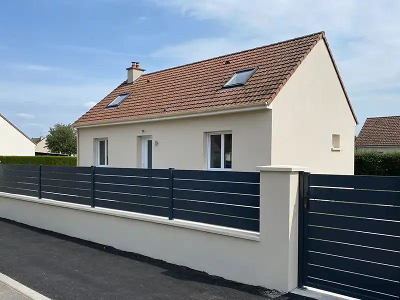 Création muret et cloture - Girault Ravalement Une petite maison moderne de couleur crème avec un toit de tuiles rouges, des fenêtres à cadre blanc et des lucarnes. La propriété est dotée d'une clôture et d'un portail en lattes horizontales gris foncé, issus d'un projet de création muret et cloture, et d'un trottoir qui longe la façade.