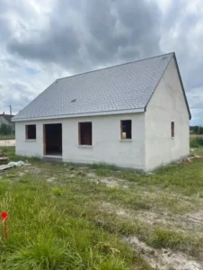 Une petite maison blanche inachevée avec un toit en bardeaux gris se dresse sur un terrain herbeux sous un ciel nuageux. Les fenêtres et la porte d'entrée ouvertes laissent apparaître des matériaux de construction à proximité, attendant la touche finale de l'enduit extérieur.
