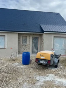 Une maison nouvellement construite avec un enduit extérieur, un toit sombre et des fenêtres couvertes, entourée de tas de gravier et de matériel de construction, dont un tonneau bleu et un compresseur d'air jaune sur une remorque à l'avant.