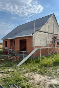 Une maison en construction avec des échafaudages sur le côté, des murs extérieurs partiellement finis en attente d'enduit extérieur, des briques apparentes, des matériaux de construction et de l'herbe au premier plan sous un ciel partiellement nuageux.