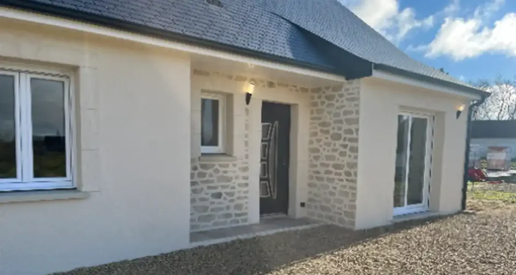 Enduit extérieur maison neuve (3) Maison de plain-pied avec une façade extérieure en pierre claire et enduit crème, un toit sombre, une porte d'entrée moderne, deux fenêtres et une cour en gravier. Le ciel est partiellement nuageux à l'arrière-plan.