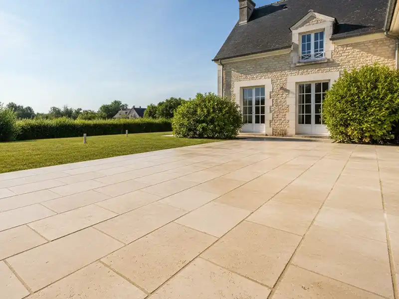 Nettoyage dallage - Girault Ravalement Un grand patio carrelé de couleur claire à l'extérieur d'une maison en pierre avec des fenêtres et des portes à cadre blanc, entouré de buissons verts et d'une pelouse bien entretenue sous un ciel bleu clair, idéal pour mettre en valeur un nettoyage dallage expert.