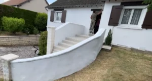 Une personne se tient debout sur un escabeau pour peindre l'habillage extérieur d'une maison blanche aux volets bruns, qui possède un escalier courbe menant à l'entrée et à un petit jardin.