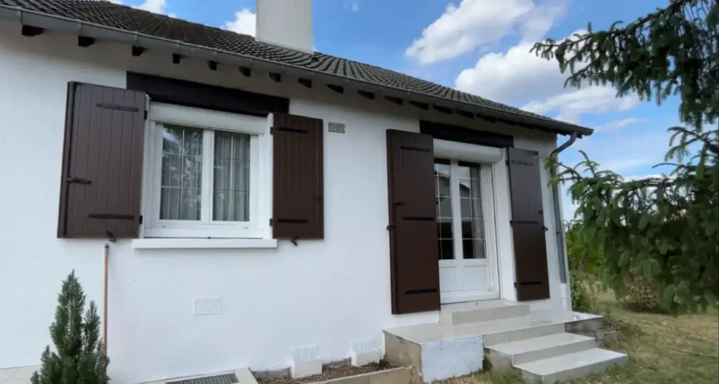 Ravalement maison individuelle 41 (3) - Girault Ravalement Une petite maison blanche avec des volets bruns, une fenêtre à gauche, une porte vitrée avec des marches à droite et de la verdure autour de l'entrée présente un ravalement maison individuelle récent sous un ciel partiellement nuageux.