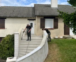 Deux personnes sont en train de nettoyer, à l'aide de nettoyeurs haute pression, les marches et la balustrade de l'entrée d'une maison. La maison a des murs blancs et beiges, des volets bruns et un toit gris en pente. Une personne se trouve sur les marches, une autre se tient sur l'herbe.