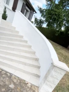 Un escalier extérieur carrelé blanc avec une rampe blanche incurvée et un escalier propre mène à une maison aux volets bruns, entourée de gazon, d'arbres et d'un sentier en pierre.