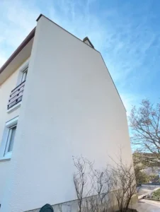 Une maison de trois étages avec un mur latéral blanc, plat et sans fenêtre, récemment mis en valeur par la rénovation de la façade, quelques arbustes nus à la base, et un ciel bleu vif au-dessus.