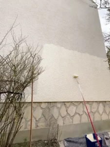 Une personne utilise un rouleau de peinture sur une perche pour appliquer de la peinture de façade sur le mur extérieur brut d'un bâtiment, créant ainsi un contraste évident entre les zones fraîchement peintes et les zones non peintes. Un buisson sans feuilles se trouve à proximité.