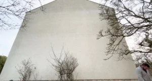 Un grand bâtiment blanc, sobre et sans fenêtres se dresse derrière des arbres et des buissons clairsemés et sans feuilles, suggérant un éventuel besoin de rénovation de façade. Dans le coin inférieur droit, une personne regarde vers le bas sous un ciel couvert.