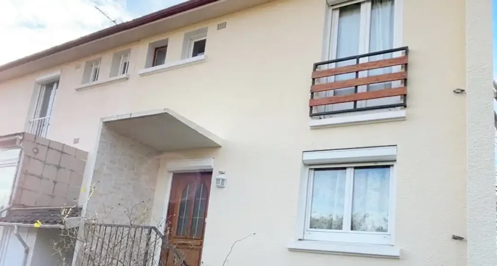 Rénovation de façade sur maison à étage - Girault Ravalement Une maison de deux étages de couleur crème avec une porte d'entrée en bois, de grandes fenêtres et un petit balcon avec des lattes en bois, avec une entrée couverte. L'extérieur simple présente des bordures blanches autour des fenêtres, magnifiquement mises en valeur par un récent ravalement de façade.
